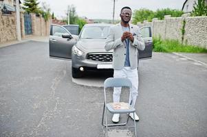 homme afro-américain riche et élégant en blazer et pantalon blanc, lunettes posées contre une voiture suv. photo