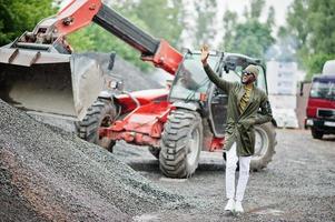 élégant homme afro-américain en chapeau et lunettes de soleil posé en plein air sous la pluie contre un tracteur avec un seau. photo