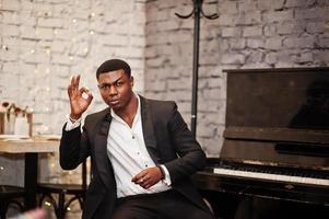 homme afro-américain puissant et fort en costume noir assis contre le piano et montrer le signe ok. photo