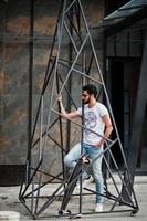 homme arabe de style rue à lunettes avec longboard posé à l'intérieur de la construction pyramidale en métal. photo