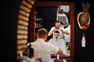 jeune homme barbu se coupe les cheveux par un coiffeur assis sur une chaise au salon de coiffure. âme de barbier. photo