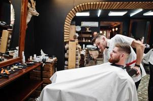 jeune homme barbu se coupe les cheveux par un coiffeur assis sur une chaise au salon de coiffure. âme de barbier. photo