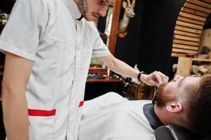 jeune homme barbu se coupe les cheveux par un coiffeur assis sur une chaise au salon de coiffure. âme de barbier. photo