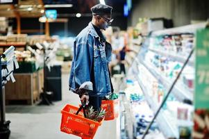 homme afro-américain décontracté et élégant à la veste en jean et au béret noir tenant deux paniers, debout près du réfrigérateur et faisant ses courses au supermarché.