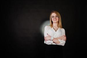 portrait en studio d'une femme d'affaires blonde à lunettes, chemisier blanc et jupe noire sur fond sombre. femme réussie et concept de fille élégante. photo