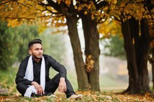 homme élégant indien en vêtements traditionnels noirs avec foulard blanc posé en plein air contre l'arbre de feuilles d'automne jaune. photo