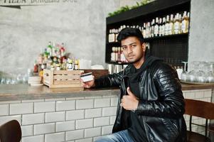jeune homme asiatique décontracté et élégant portant une veste en cuir assis sur un bar et tenant une carte de crédit pour payer. photo