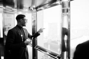 homme afro-américain à la mode en costume et lunettes avec téléphone portable et tasse de café aux mains posées à l'intérieur de l'ascenseur. photo