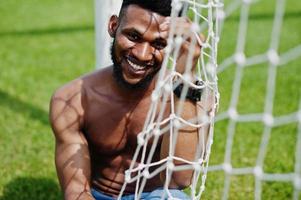 bel homme musclé torse nu afro-américain sexy à la salopette en jean posée à l'herbe verte contre les portes de football du terrain du stade. portrait d'homme noir à la mode. photo