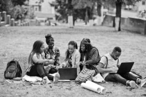 groupe de cinq étudiants africains qui passent du temps ensemble sur le campus de la cour universitaire. amis afro noirs assis sur l'herbe et étudiant avec des ordinateurs portables. photo