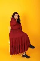 jolie femme sud-asiatique en robe rouge foncé posée au studio sur fond jaune et assise sur une chaise. photo