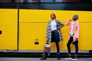 deux jeunes femmes musulmanes africaines modernes à la mode, attrayantes, grandes et minces en hijab ou turban foulard et manteau posés contre un bus jaune.