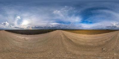 panorama hdri sphérique complet et harmonieux vue à 360 degrés sur la route de gravier parmi les champs au printemps avec des nuages impressionnants en projection équirectangulaire, prêt pour le contenu de réalité virtuelle vr ar photo