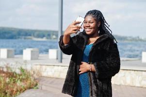 modèle de taille plus afro-américain à la peau foncée posé dans une robe bleue brillante et un manteau de fourrure noir contre le bord de la mer faisant selfie au téléphone. photo