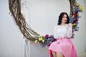 jeune fille brune en jupe rose et chemisier blanc posé à l'intérieur contre une grande couronne décorée. photo