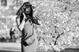 élégante fille afro-américaine en tunique grise, sac à bandoulière et casquette posée à la journée d'automne ensoleillée contre les feuilles jaunes. femme modèle afrique. photo