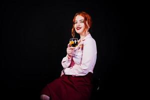 magnifique fille aux cheveux rouges en chemisier rose et jupe rouge avec un verre de vin à portée de main isolé sur noir. photo