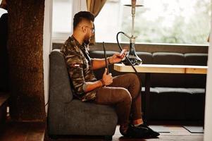 homme arabe à barbe élégante dans des verres et une veste militaire fumant le narguilé au bar de la rue. modèle arabe se reposant et prenant un selfie au téléphone. photo