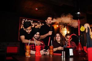 groupe d'amis indiens s'amusant et se reposant dans une boîte de nuit, buvant des cocktails et fumant du narguilé, regardant des téléphones portables. photo