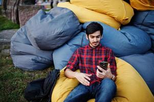 jeune étudiant indien en chemise à carreaux et jeans assis et se détendre sur des oreillers extérieurs. passer du temps avec le téléphone portable. photo