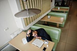 un homme arabe porte une veste en jean noir et des lunettes assis dans un café, lit un livre et boit du café. mec modèle arabe élégant et à la mode. photo