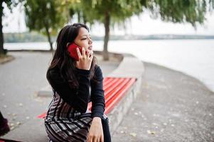 portrait d'une jeune belle adolescente indienne ou sud-asiatique en robe assise sur un banc avec un téléphone portable. photo