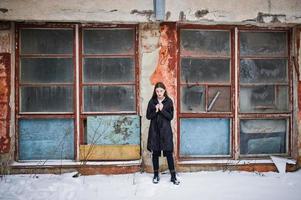 modèle brune à longues jambes à la mode dans un long manteau noir posé en plein air le jour de l'hiver contre le vieux mur de grunge avec des fenêtres cassées. photo