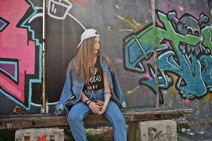 une fille hipster décontractée et élégante en casquette et en jean porte de la musique à partir d'un casque de téléphone portable contre un grand mur de graffitis avec une bombe. photo