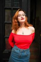 jolie femme rousse à lunettes, porter un chemisier rouge et une jupe en jean posant dans la rue contre la vieille porte en bois. photo