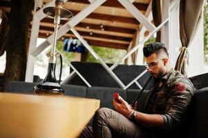 homme arabe à barbe élégante dans des verres et une veste militaire fumant le narguilé au bar de la rue. modèle arabe se reposant et regardant au téléphone. photo