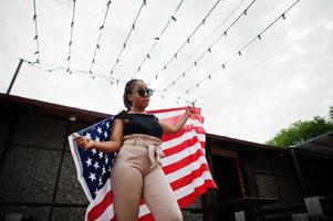 élégante femme afro-américaine à lunettes de soleil posée en plein air avec le drapeau américain. photo