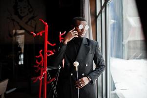 élégant gentleman afro-américain dans une élégante veste noire et des lunettes, tenant un bâton de marche rétro comme un flacon de canne ou une canne basculante avec une poignée en boule de diamant doré et parlant au téléphone mobile. photo