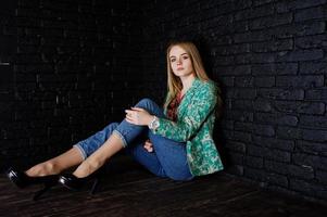 fille blonde élégante en veste et jeans contre le mur noir brique au studio. photo