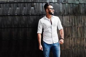 grand modèle d'homme arabe élégant en chemise blanche, jeans et lunettes de soleil posés contre un mur en bois à l'intérieur. Barbe mec arabe attrayant. photo