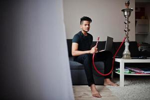 homme indien beau et à la mode en noir assis dans la chambre, fumant du narguilé et travaillant sur un ordinateur portable. photo