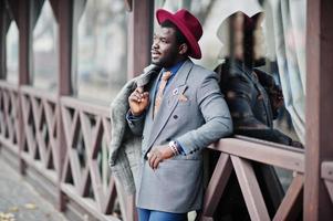 élégant modèle d'homme afro-américain en manteau gris, cravate de veste et chapeau rouge posé contre un café en bois. photo