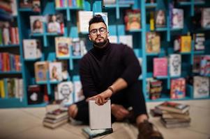 grand étudiant arabe intelligent, portant un col roulé violet et des lunettes, à la bibliothèque assis contre des étagères de livres. photo