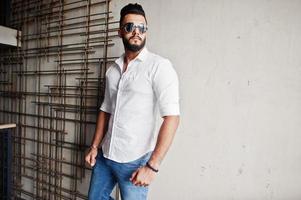 grand modèle d'homme arabe élégant en chemise blanche, jeans et lunettes de soleil posés contre un mur d'acier à l'intérieur. Barbe mec arabe attrayant. photo