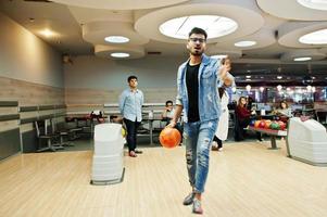 homme asiatique élégant en veste de jeans et lunettes debout au bowling avec balle à portée de main et lancez-le. photo