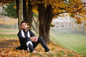 homme élégant indien en vêtements traditionnels noirs avec foulard blanc posé en plein air contre l'arbre de feuilles d'automne jaune. photo