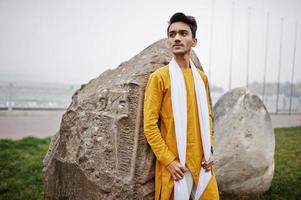 homme élégant indien en vêtements traditionnels jaunes avec foulard blanc posé en plein air contre de grosses pierres. photo