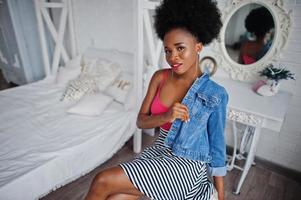 jolie femme afro-américaine aux cheveux afro portant une jupe et une veste en jean, posée dans la salle blanche. modèle noir à la mode. photo