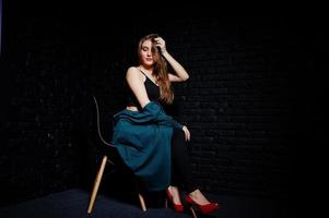 belle fille brune porte une veste noire et verte en talons hauts rouges, assise et posant sur une chaise au studio contre un mur de briques sombres. portrait de modèle de studio. photo
