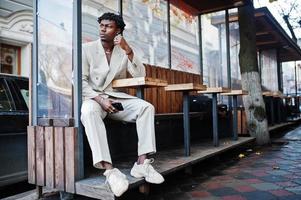 homme afro élégant en costume beige old school assis à table dans la rue. jeune homme africain à la mode en veste décontractée sur torse nu. photo