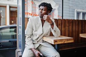 homme afro élégant en costume beige old school assis à table dans la rue. jeune homme africain à la mode en veste décontractée sur torse nu. photo