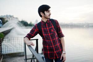 jeune étudiant indien en chemise à carreaux et jeans avec sac à dos posé sur la ville du soir contre les fontaines. photo