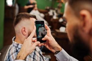 bel homme barbu au salon de coiffure, coiffeur au travail, faisant des photos sur son téléphone.