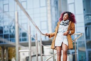 jolie femme afro-américaine bouclée en manteau marron posée près des garde-corps contre un bâtiment moderne à plusieurs étages. photo