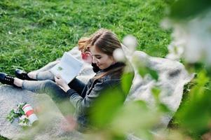 jeune fille brune en jeans assis sur un plaid contre un arbre de fleurs de printemps et lire le livre. photo