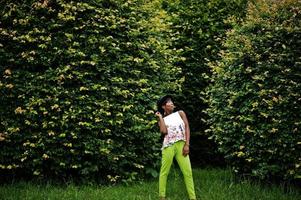 incroyable femme modèle afro-américaine en pantalon vert et chapeau noir posé au parc. photo